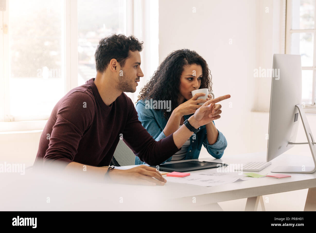 Businessman pointing towards the computer while discussing work with a business partner. Woman entrepreneur drinking coffee while discussing work with Stock Photo