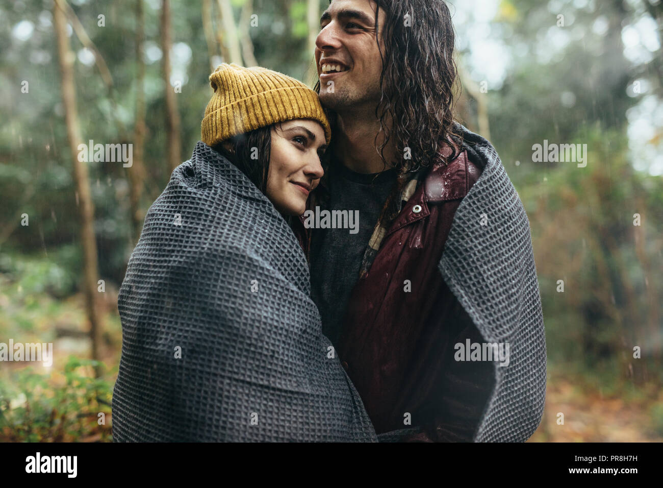Beautiful young couple wrapped in a blanket on a rainy day. Caucasian couple embracing under the rain. Stock Photo
