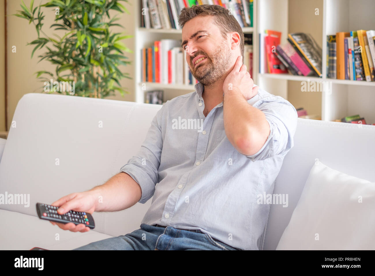 Man suffering neck ache seated on the sofa watching television with bad
