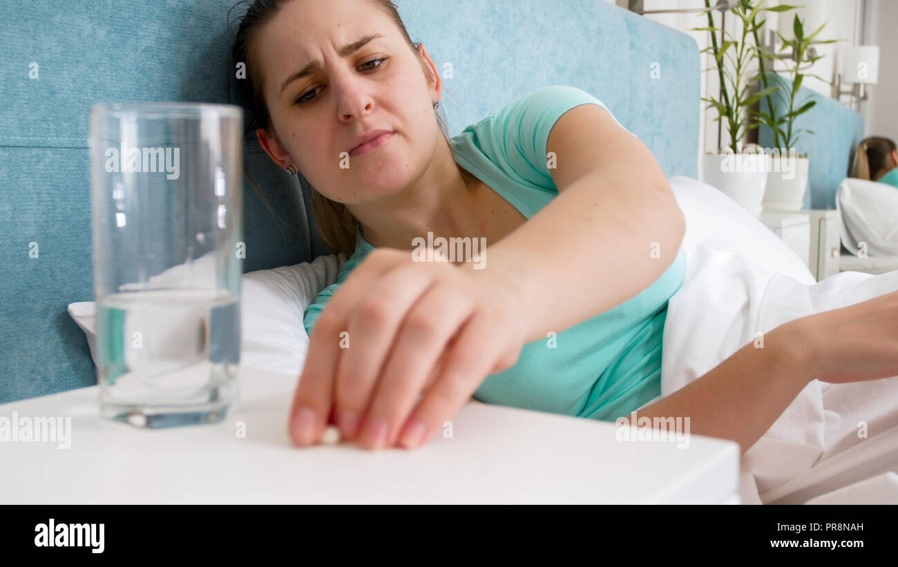 Portrait Of Young Sick Woman Lying In Bed And Taking Pill From Bedside Table Portrait Of Young Sick Woman Lying In Bed And Taking Pill From Bedside Table