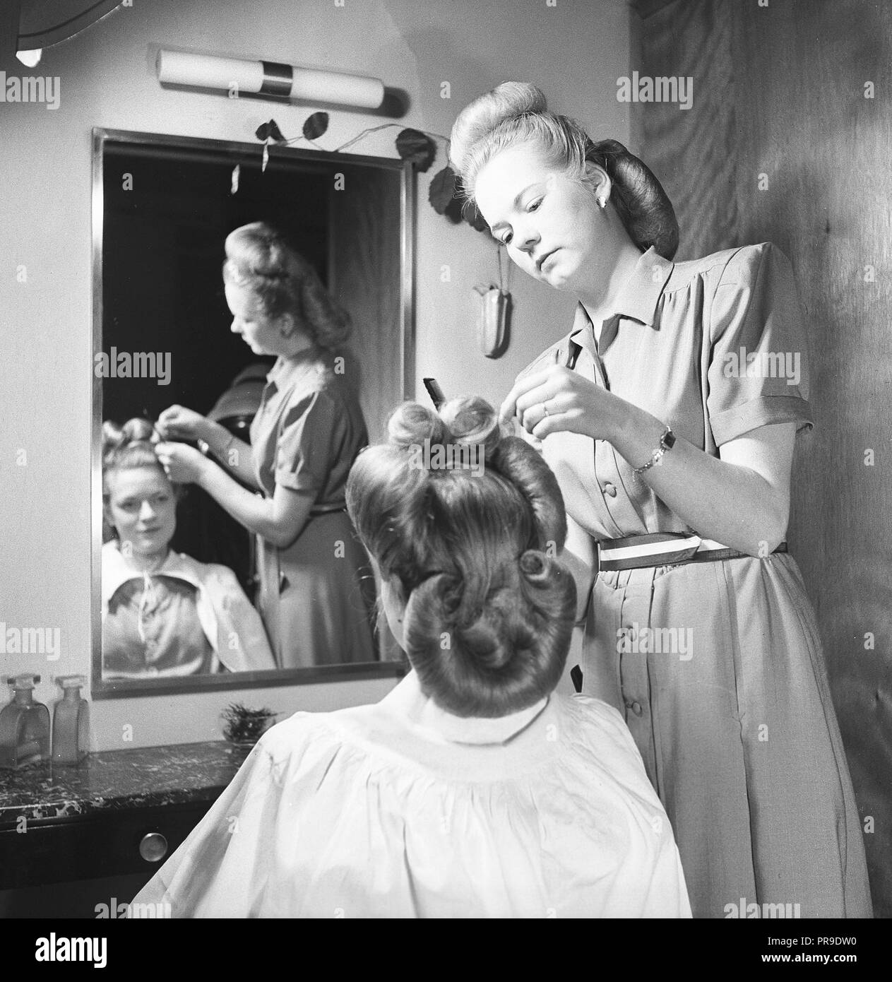 Getting a 1940s hairdo.  A dark haired young woman is looking at herself in the mirror at the ladies hairdresser's salon while a hairdresser is styling and shaping the hair into a typical 1940s hairdo. Sweden 1947. Photo Kristoffersson ref AB22-7 Stock Photo