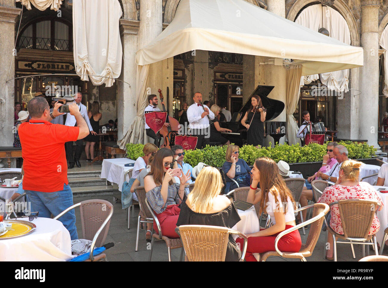 Man takes photo of orchestra playing for customers in front of iconic Florian cafe in St Mark's Square, Venice, Italy. Image shows musicians playing. Stock Photo