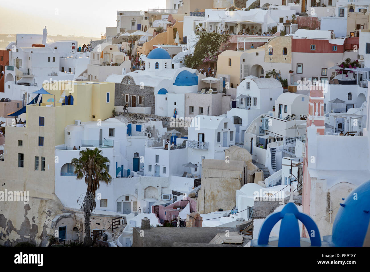 traditional cliff side houses of  Oia landmark blue dome churches Santorini, a Cyclades group of islands in Greece, tourists walking up the steep hill Stock Photo