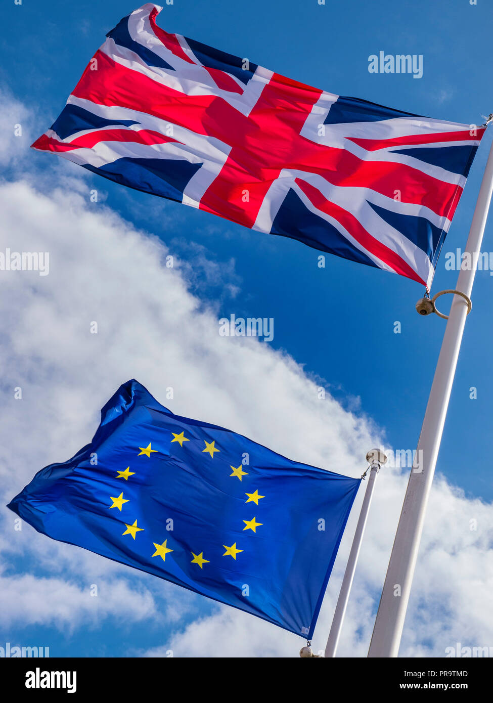 BREXIT CONCEPT FLAGS UK Union Jack Flag flying high above EU European Flag in a stiff breeze on a sunlit day with blue sky divided by clouds Stock Photo