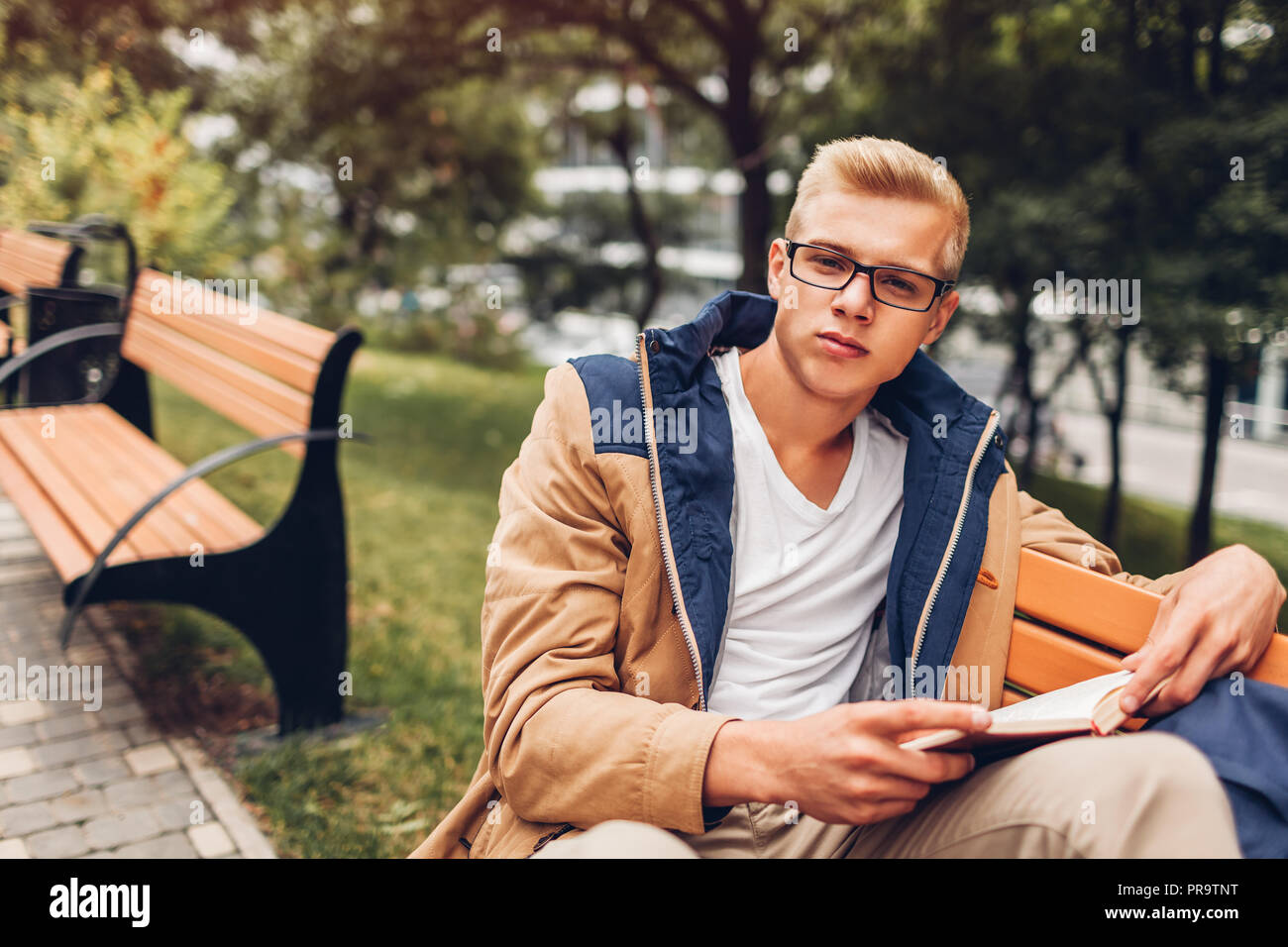 College student with backpack reading book walking in autumn park sitting on bench. Man studying