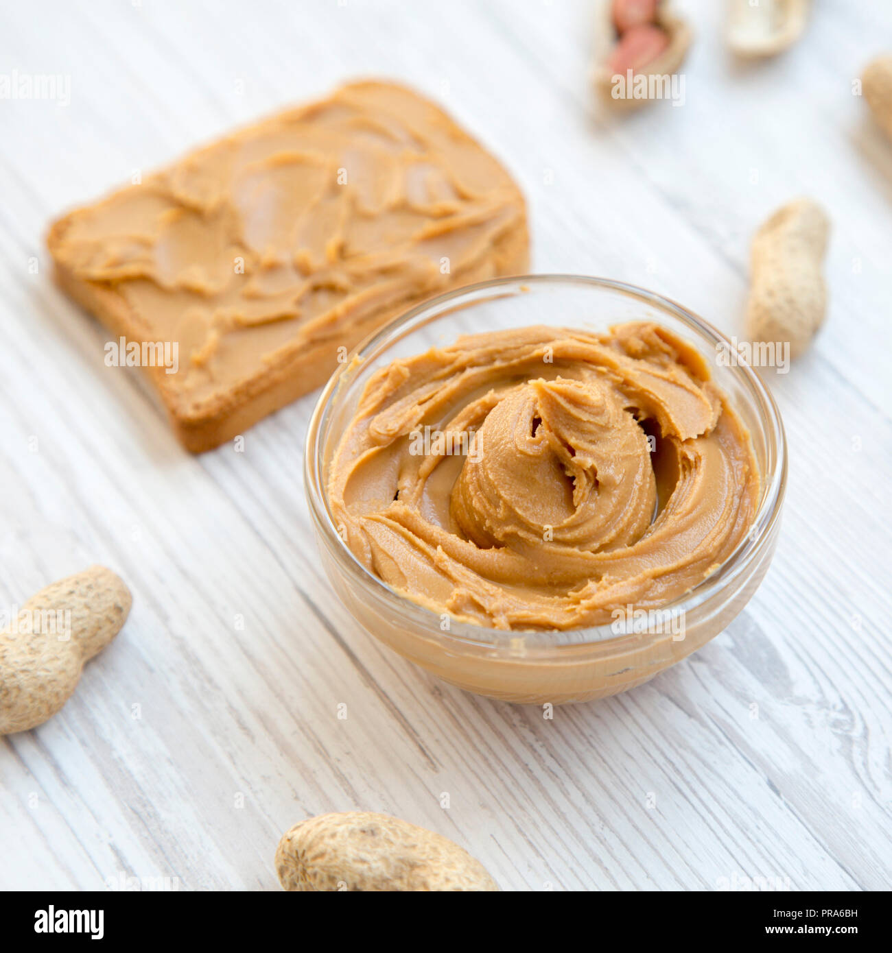 Toast, bowl of peanut butter and peanuts in shells on a white wooden table, side view. Close-up. Stock Photo