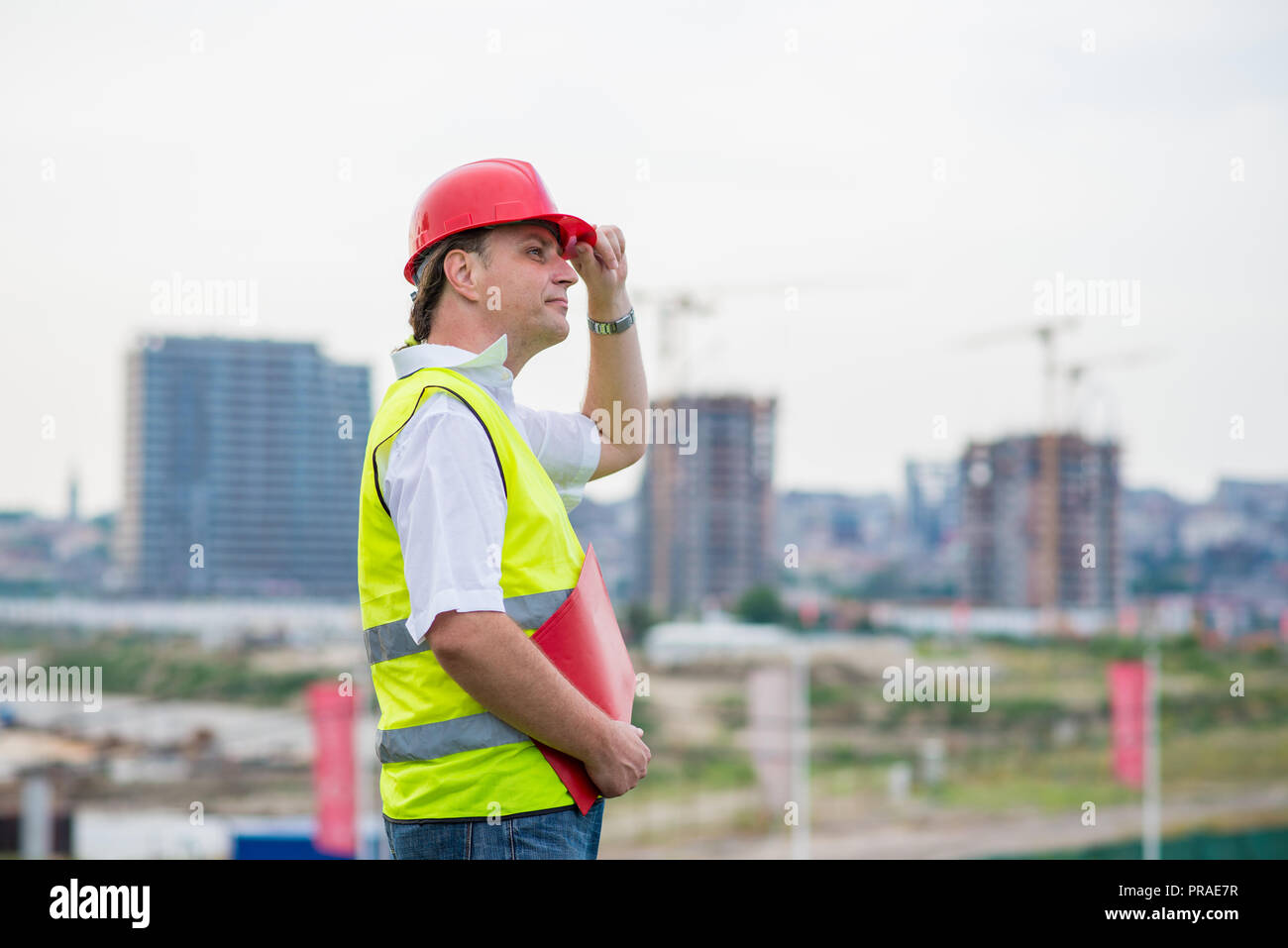 Engineer on a construction site wearing yellow reflective west and red helmet with buildings and cranes in the background. Foreman on a building site Stock Photo