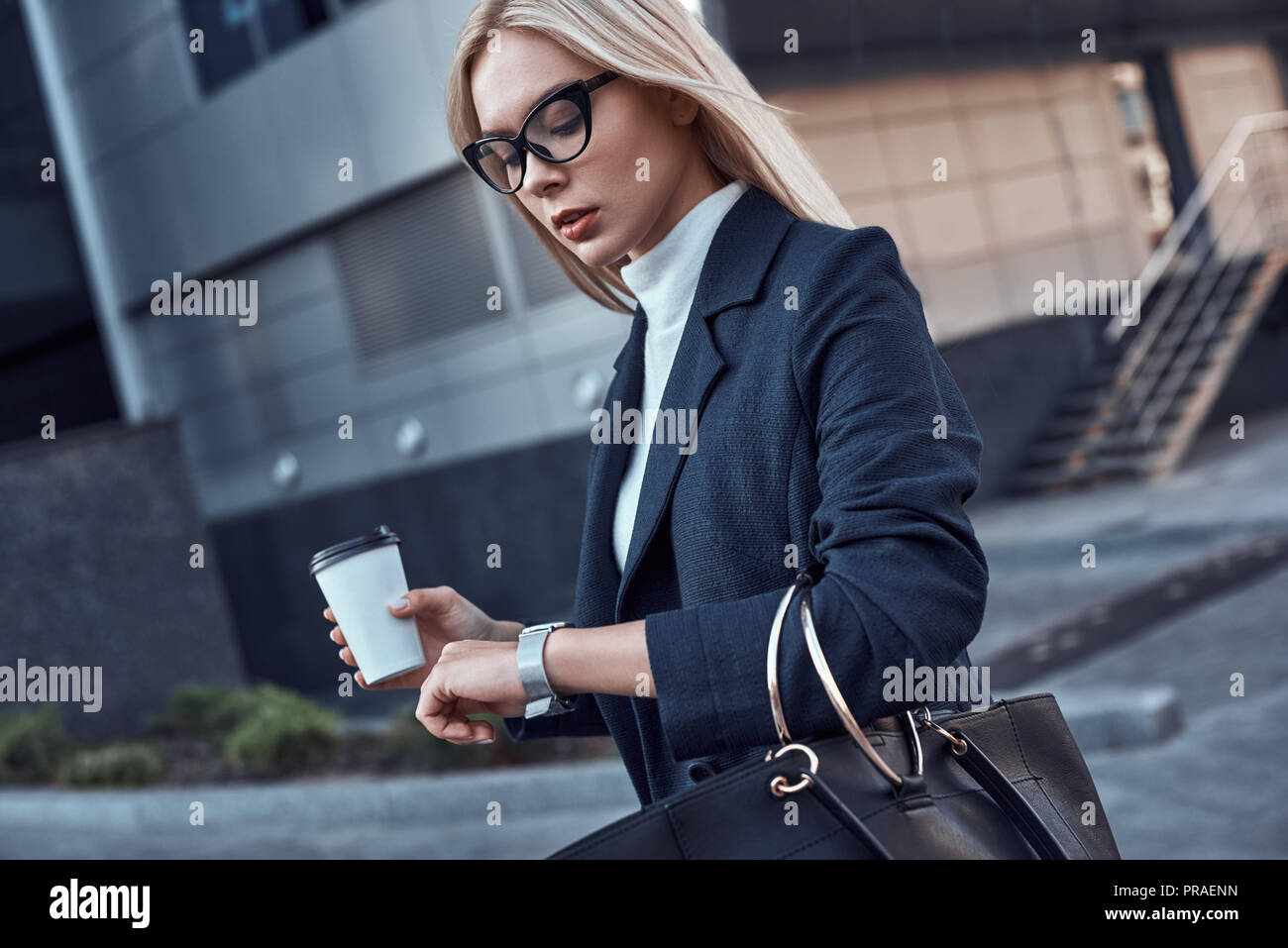 Young woman wathes at her wrist watch. In one her hand coffe, in another handbag Stock Photo