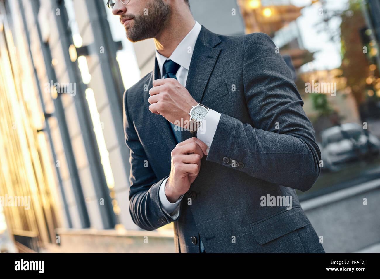 Cropped profile portrait of a successful young bearded guy in suit and glasses. So stylish and nerdy. Outdoors on a sunny street, fixing his cuffs Stock Photo