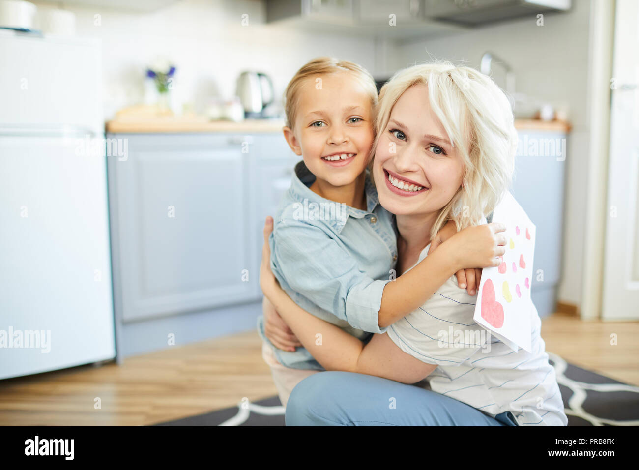 Cheerful little girls and her pretty blonde mother in embrace looking at you in the kitchen Stock Photo