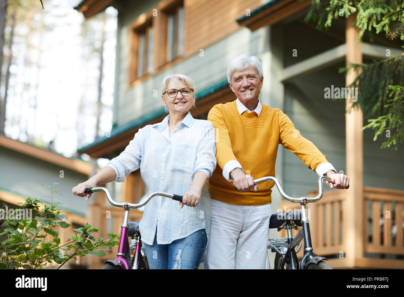 Cheerful active senior couples in casual clothing standing against country house and holding bicycles while looking at camera Stock Photo