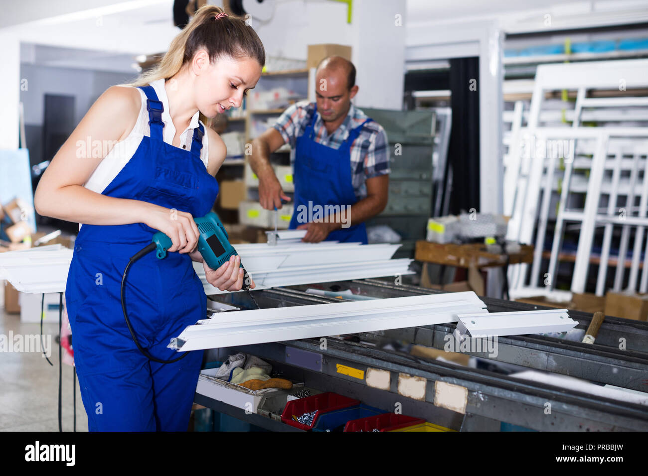 Female worker is assembling metal-plastic window in workshop, using electric drill. Stock Photo