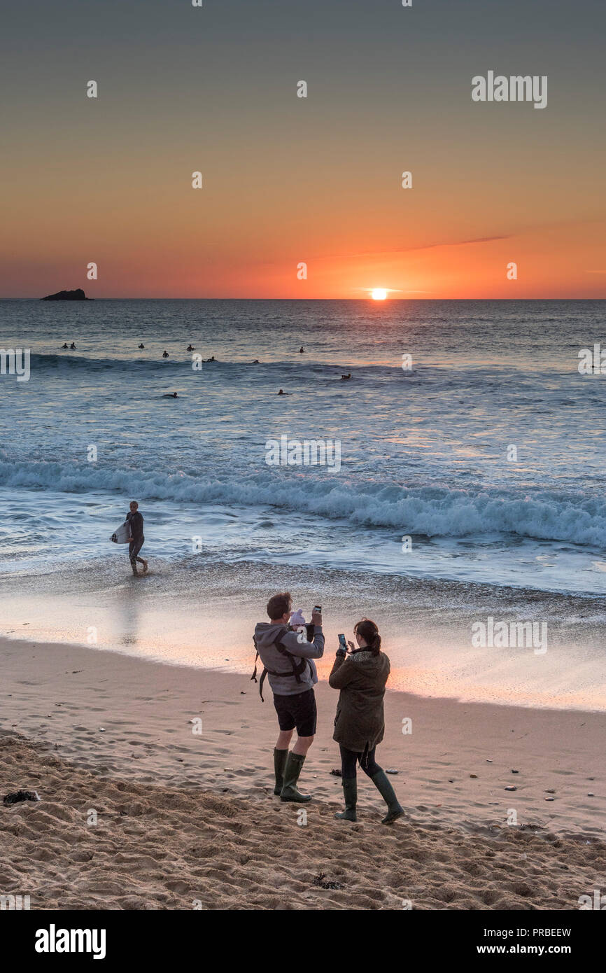 A couple and baby standing on the shore using their smartphones to photograph the intense sunset over Fistral Beach in Newquay in Cornwall. Stock Photo