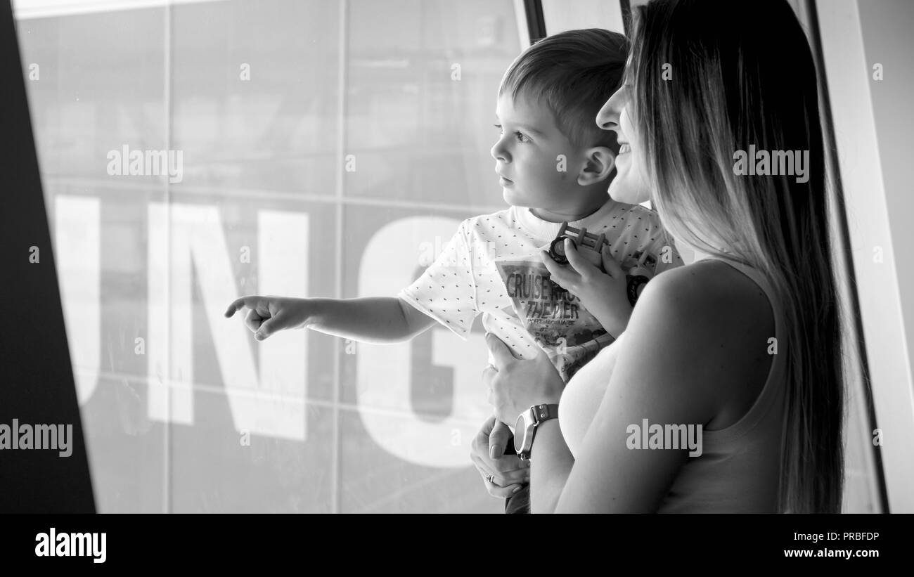 Black and white portrait of little toddler boy with mother looking out of big window at airport Stock Photo