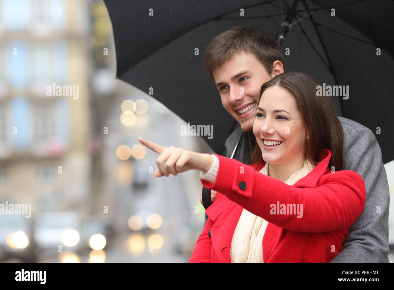 Happy couple pointing away holding an umbrella under the rain in winter in the street Stock Photo