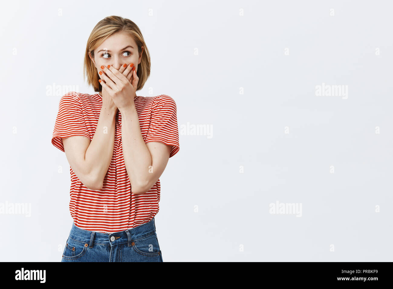 Girl witnessing embarrassing scene, losing speech, gasping, covering mouth with palms and staring anxiously right, being shocked and speechless, standing intense and scared over gray background Stock Photo