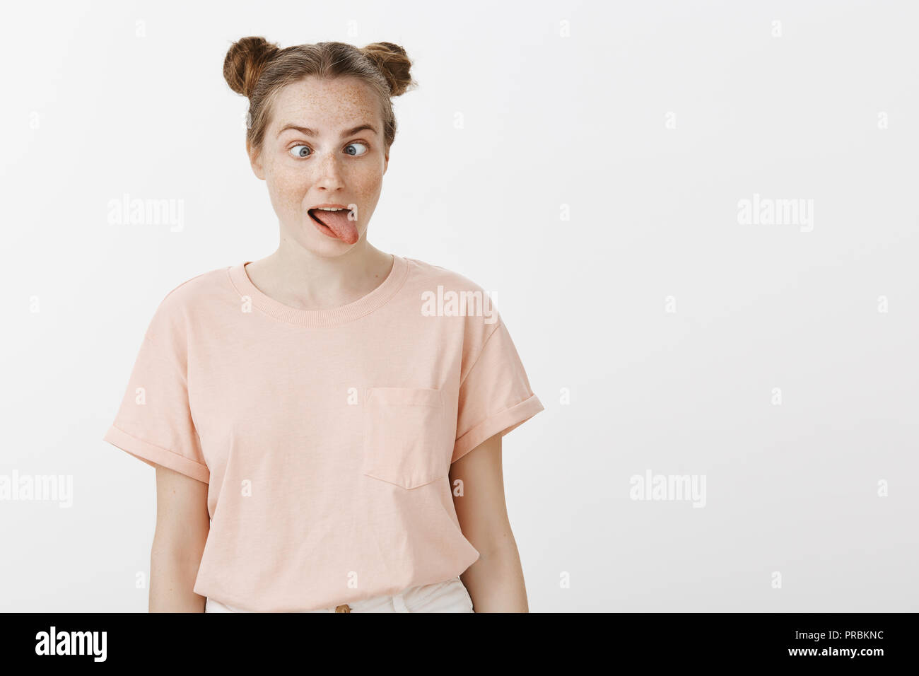 Playful bored girl with buns hairstyle and freckles in trendy pink t-shirt, sticking out tongue and squinting, being childish while having nothing to do, expressing joyful lifestyle over grey wall Stock Photo