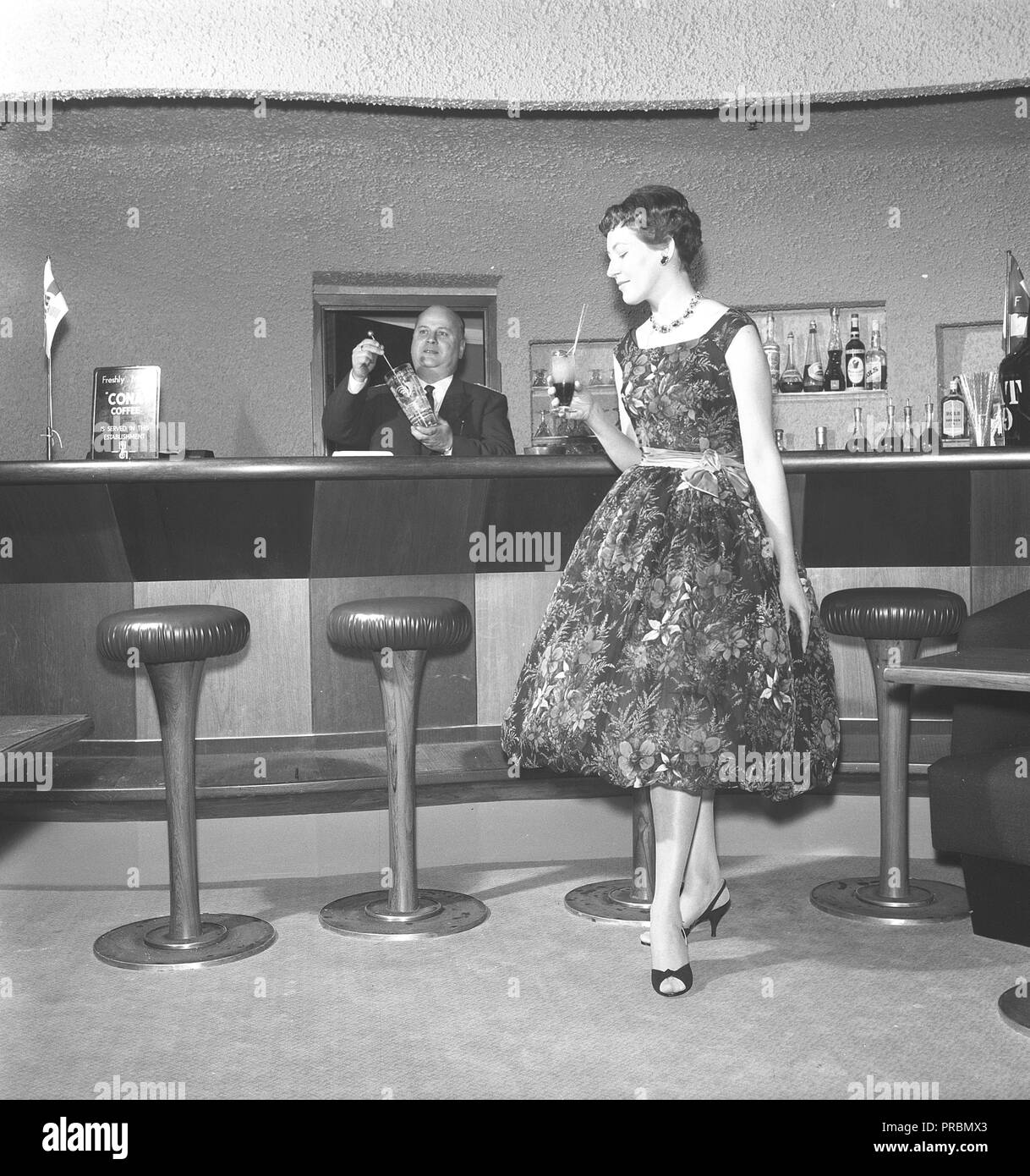 Woman in the 1950s. A young woman is standing in a bar with a drink in her hand. She is wearing a fashionable dress with flowers. Sweden 1956. Photo Kristoffersson Ref BY71-5 Stock Photo