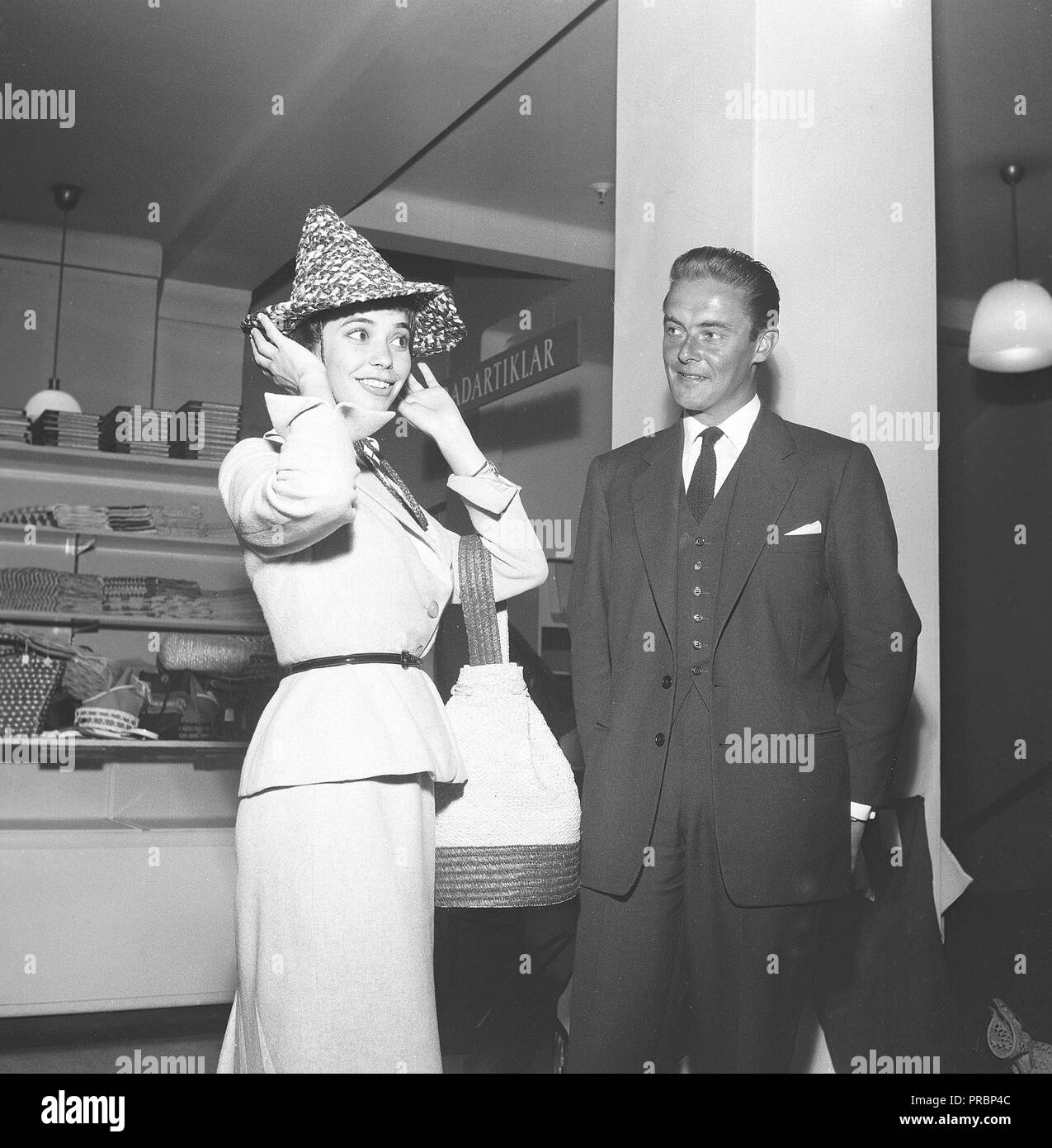1950s fashion. Swedish actress Margit Carlqvist, born 1932 is trying on a hat while a man is standing beside her looking at her. Sweden 1956. Ref BX34-6 Stock Photo