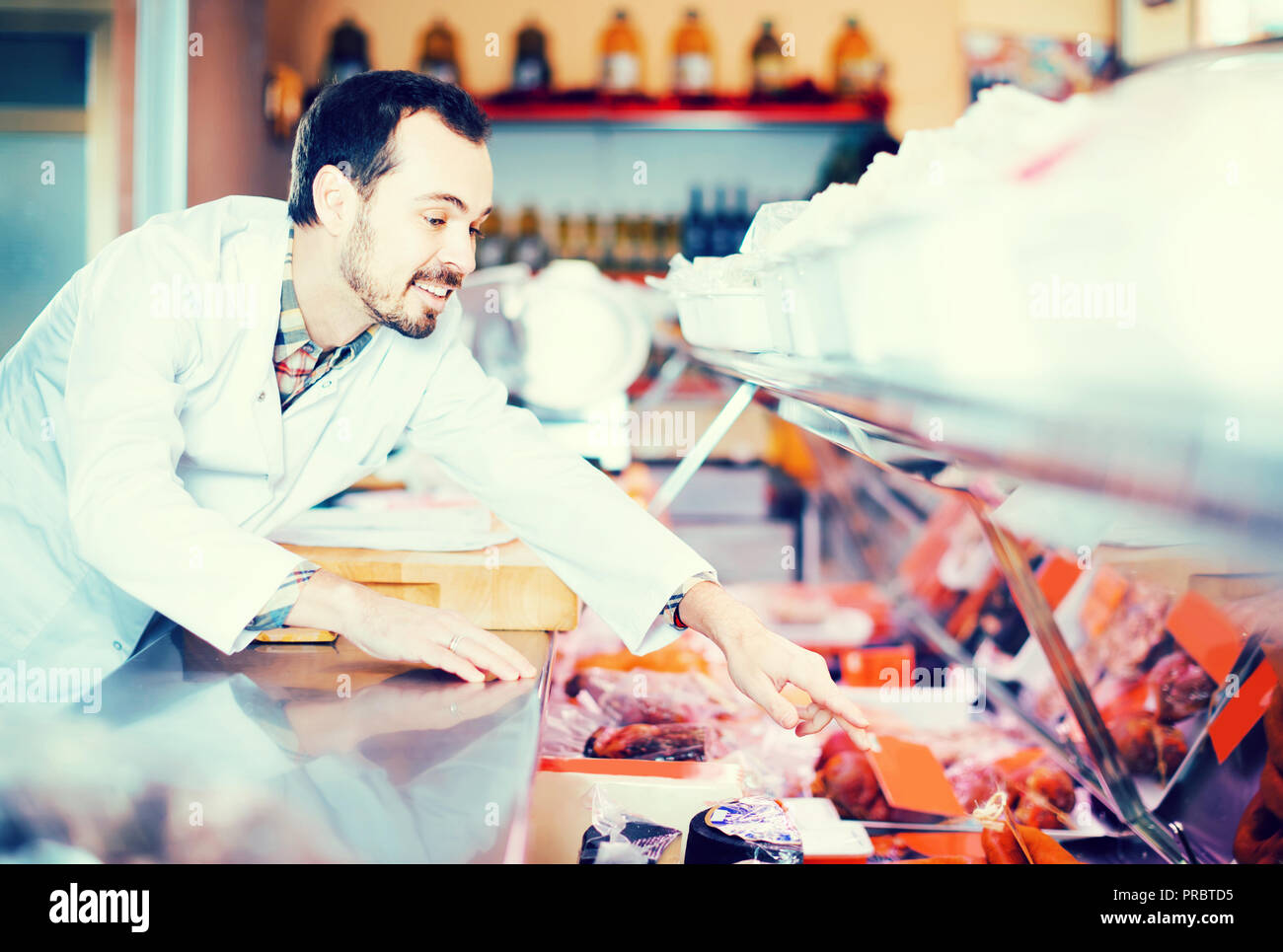 Happy Spanish Male Shop Assistant Demonstrating Sliced Bacon In Butcher