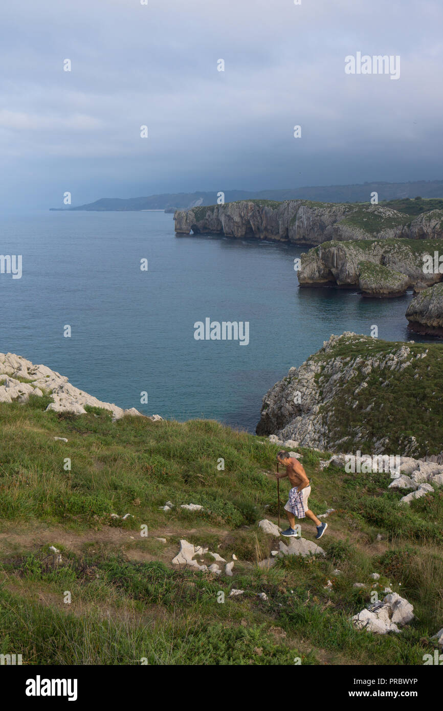 Man walking on a coastal path along rugged coastline near Playa de Cuevas del Mar, Llanes, Asturias, Spain with northern Spain coastline in background Stock Photo
