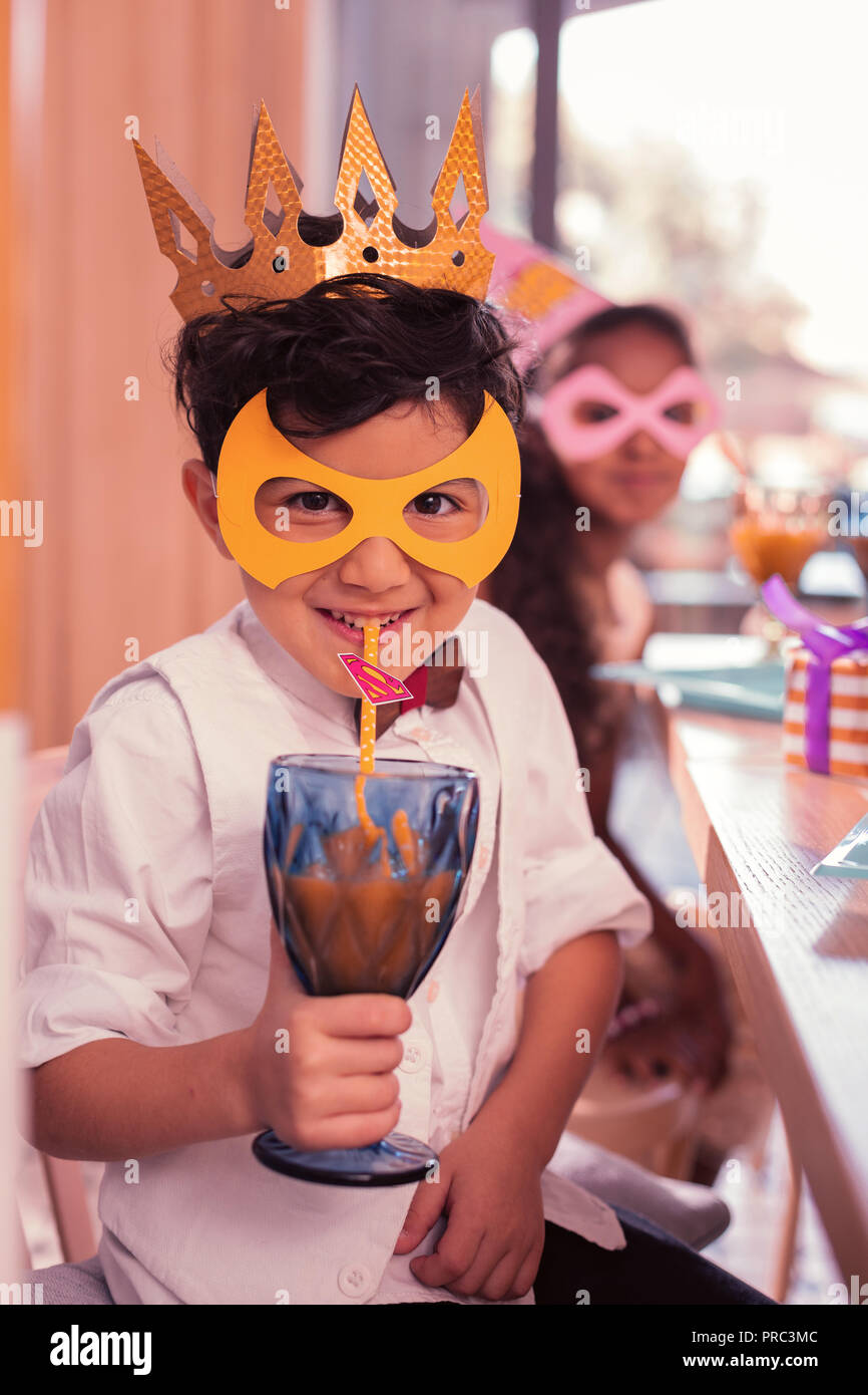 Funny boy wearing mask and smiling while drinking juice Stock Photo