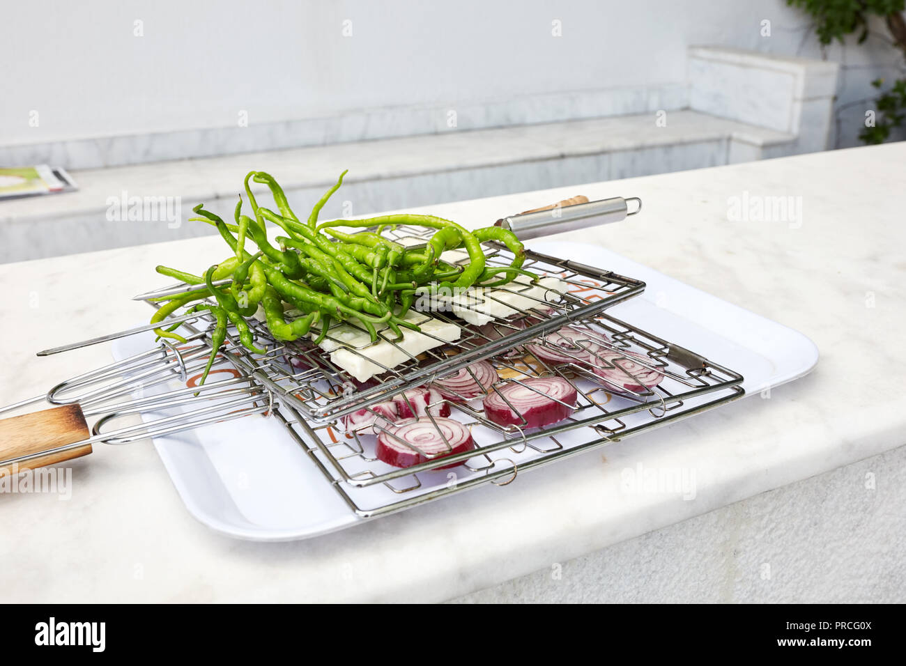 Fresh long green peppers, white cheese and sliced red onions placed between the wire grill grid on a tray over a counter and waiting to get barbecued. Stock Photo