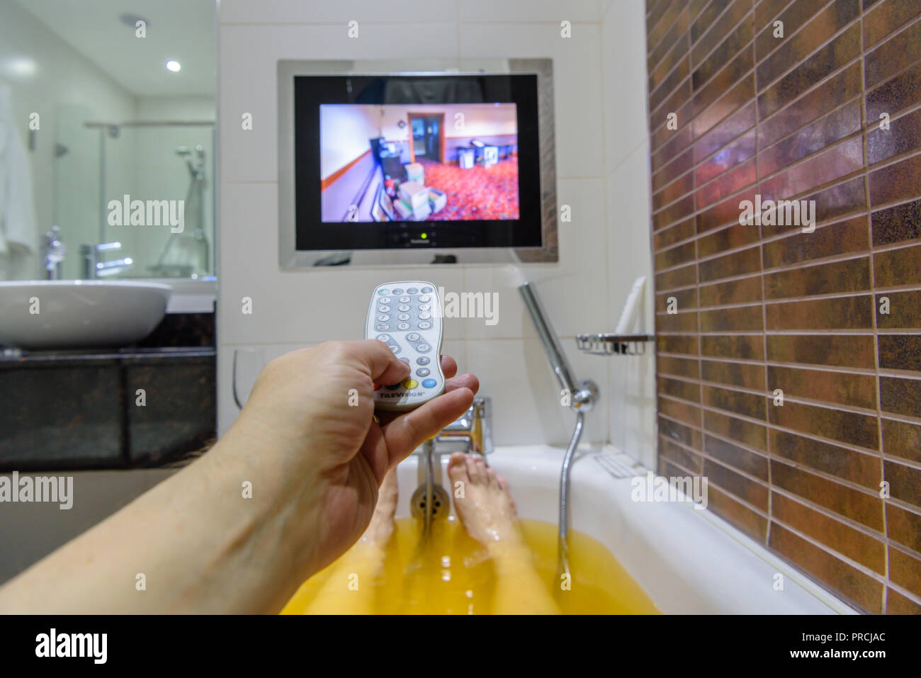 A man uses a remote control for a wall-mounted television as he takes a bath. Stock Photo