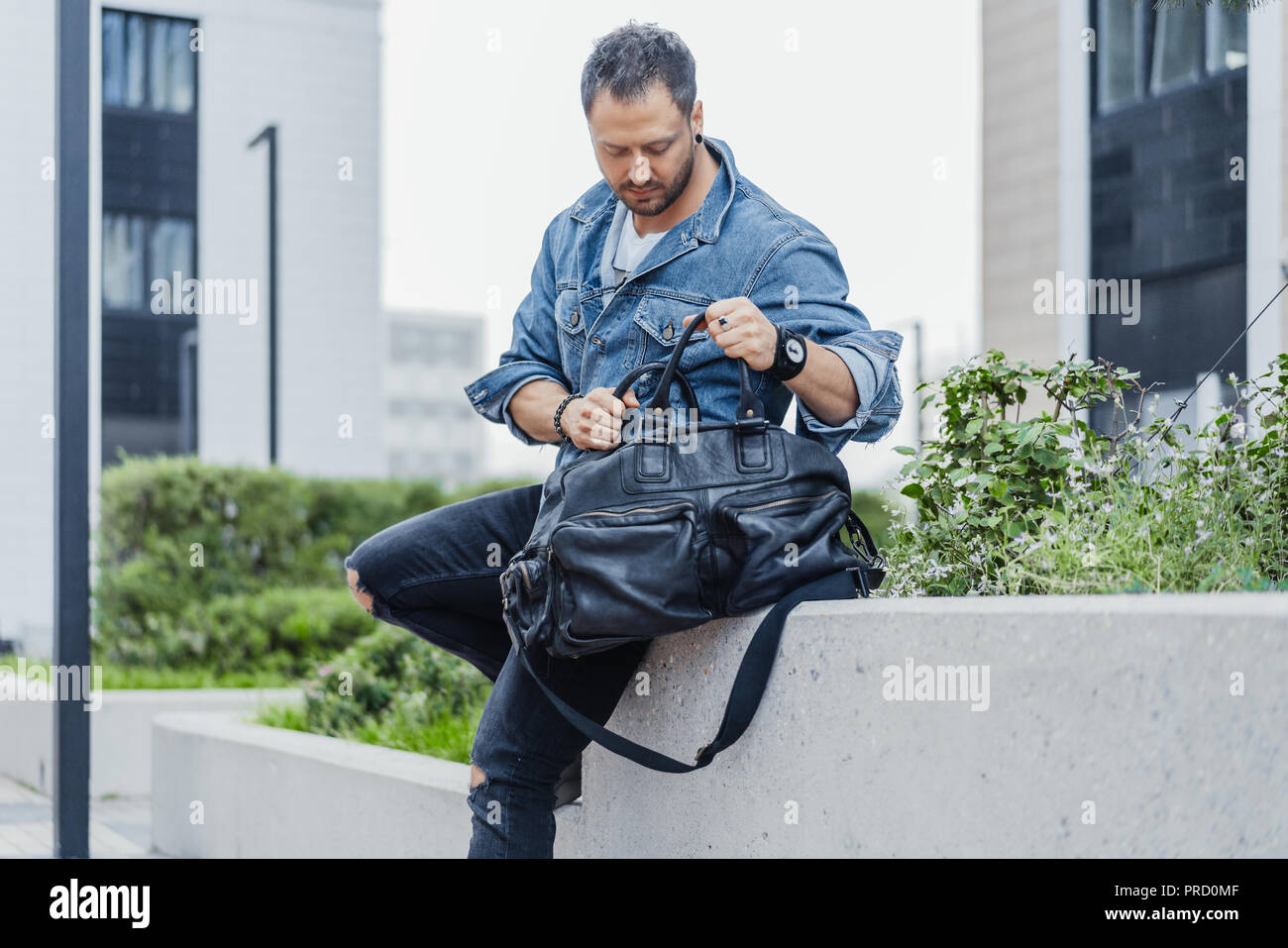 Young man rummaging in black sport bag and sitting on cityscape ...