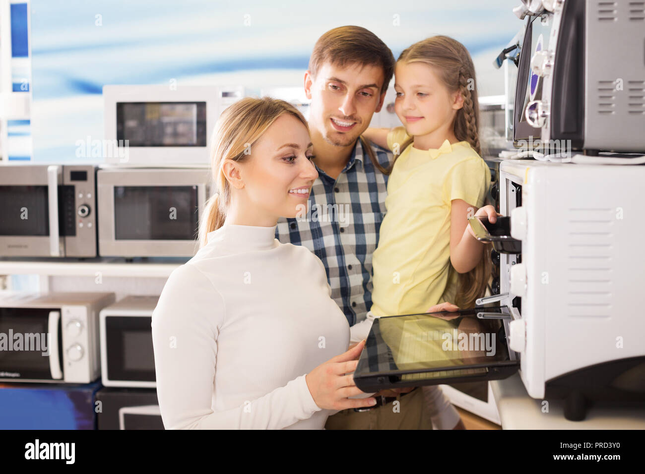 Smiling young man and woman with girl buying modern microwave in store with electronics Stock Photo