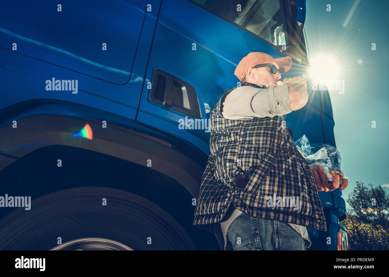Semi Truck Driver Lunch Brake in the Sun. Caucasian Trucker Enjoying His Meal in Front of His Vehicle. Stock Photo