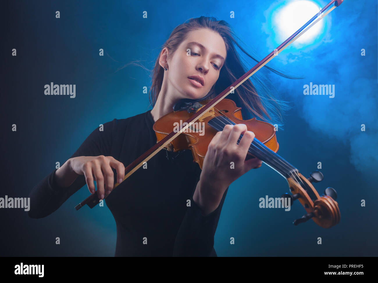 Beautiful young woman playing the violin on dark blue background. Fog in the background. Studio shot Stock Photo