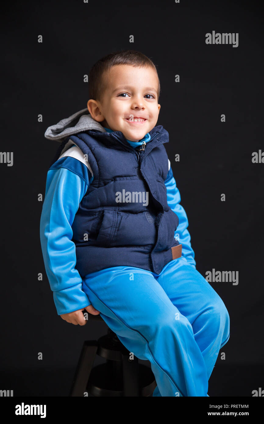 Portrait of little boy smiling and sitting on a black stool, isolated on black background Stock Photo