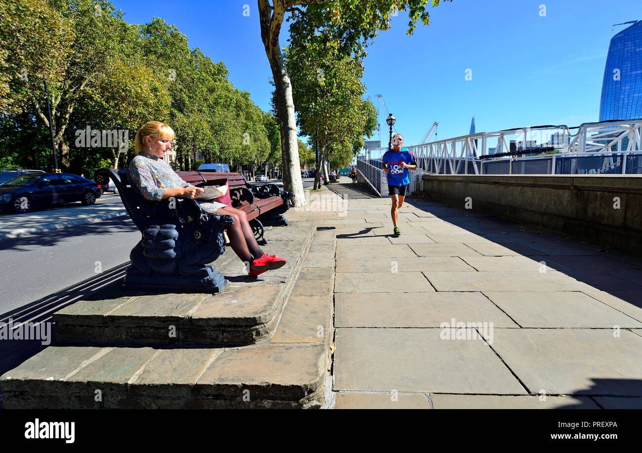Woman sitting reading as a jogger runs past on the Victoria Embankment, London, England, UK. Stock Photo