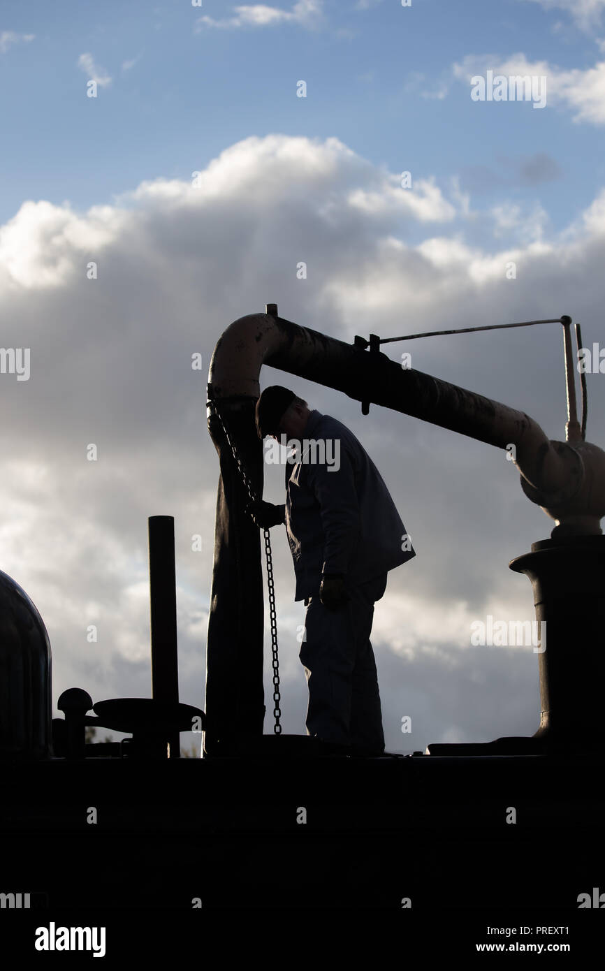 Close up of railway steam train crew, in sky silhouette, standing on vintage UK locomotive at water stop, engine taking on water, early evening. Stock Photo