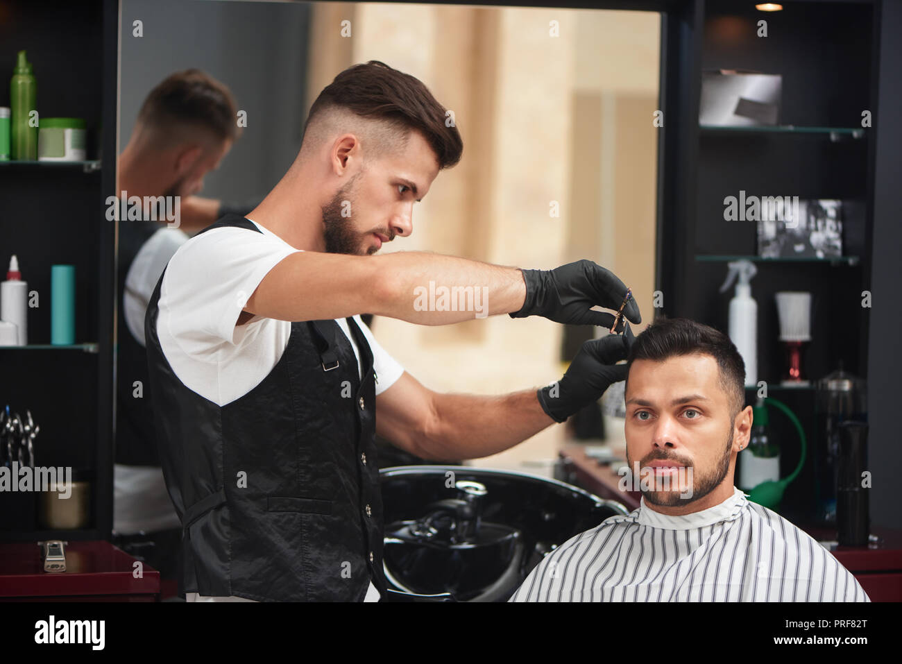 Serious male hairdresser in uniform and black gloves working in barbershop. Professional master cutting hair of man using scissors and comb. Client sitting and looking away. Stock Photo