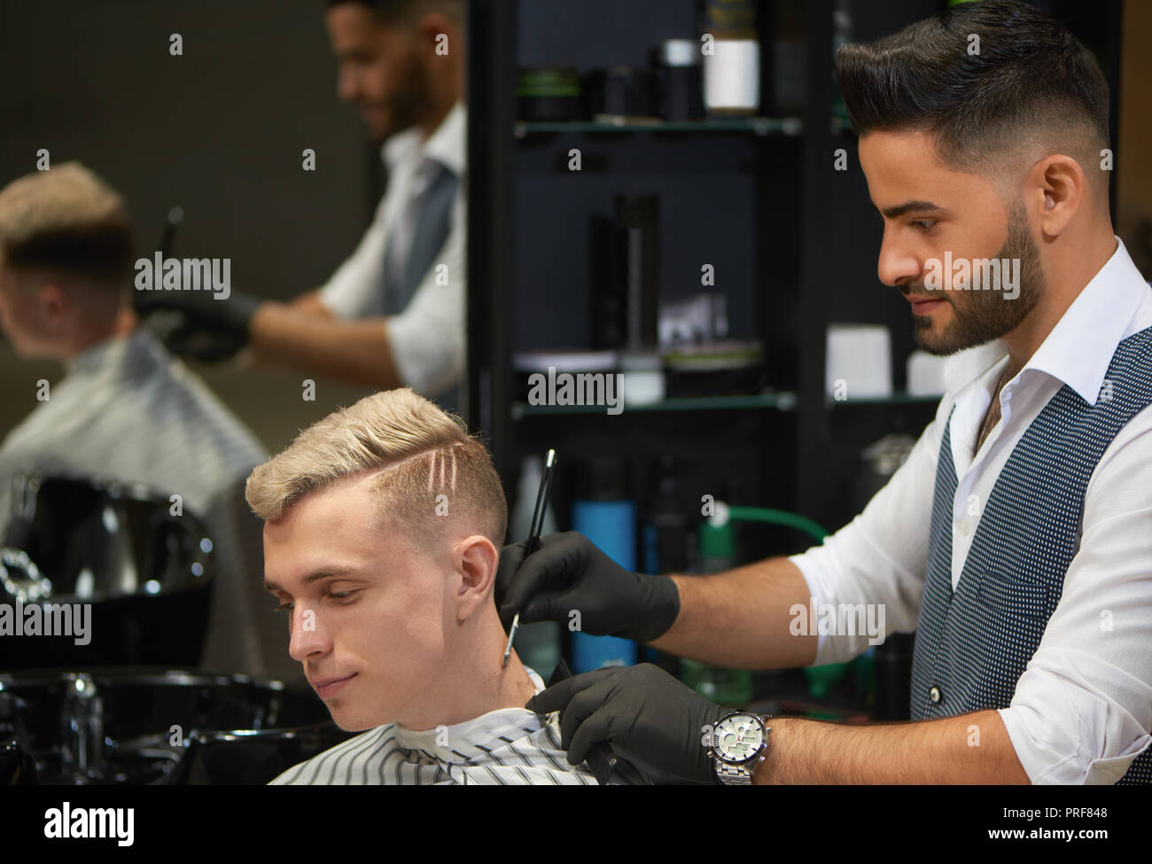 Side view of professional hairstylist shaving with razor neck of client. Handsome young man with blonde hair looking down. Bearded barber wearing white shirt, vest and black gloves. Stock Photo