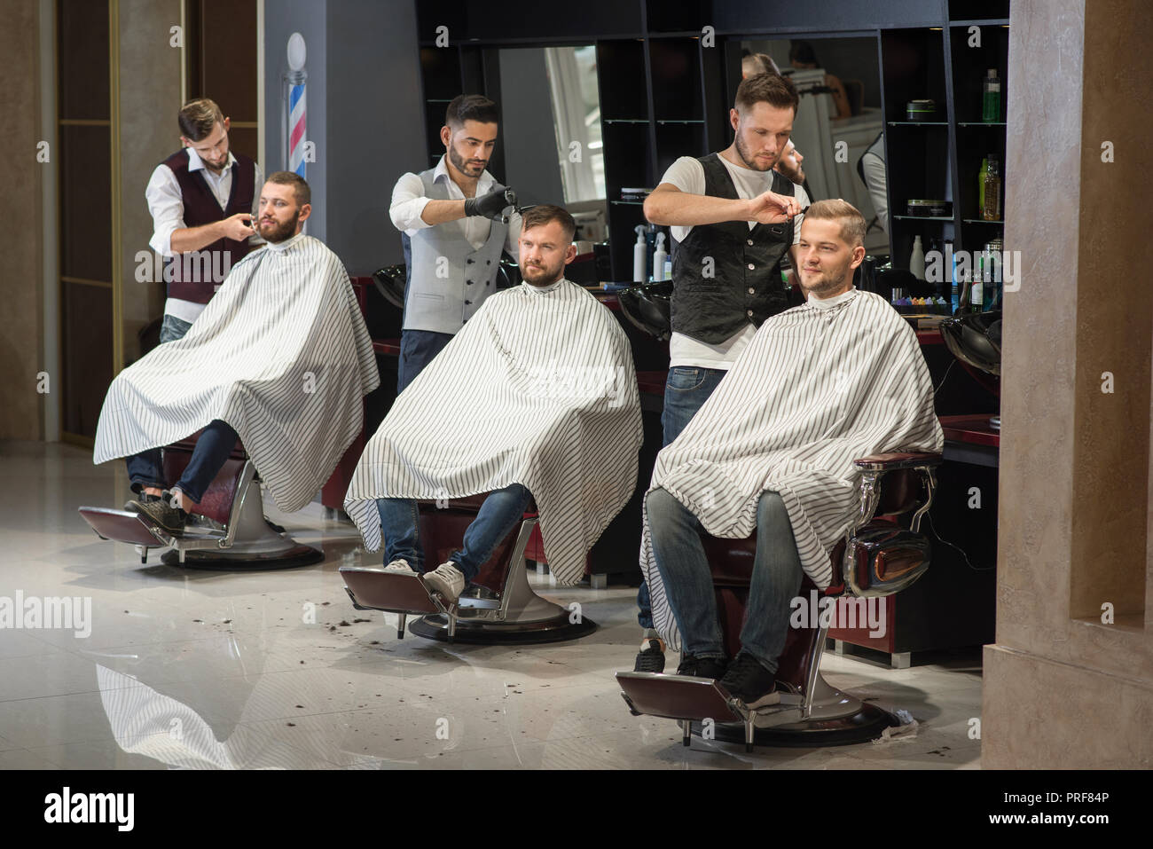 Process of styling and grooming men's haircuts in barbershop. Three professional and confident barbers standing and cutting hair of men. Male clients sitting in chairs and wearing haircut gowns. Stock Photo