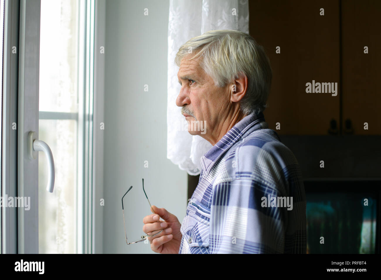 portrait of a gray-haired man 60 years old. A man looks out the window. The man has poor eyesight, he wears glasses Stock Photo