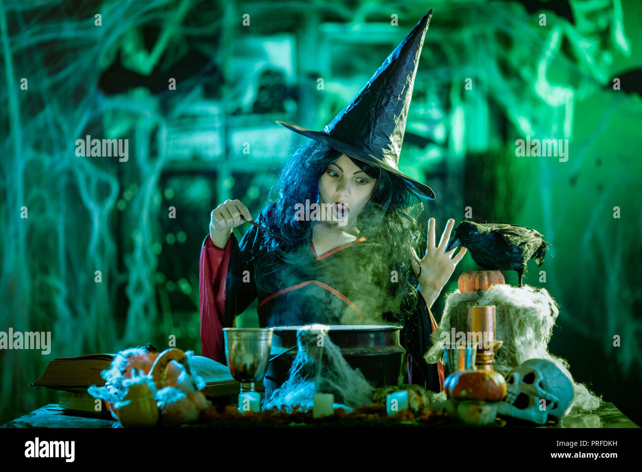 Young witch with awfully face reading recipes of magic drink in creepy surroundings and smoky background. Stock Photo