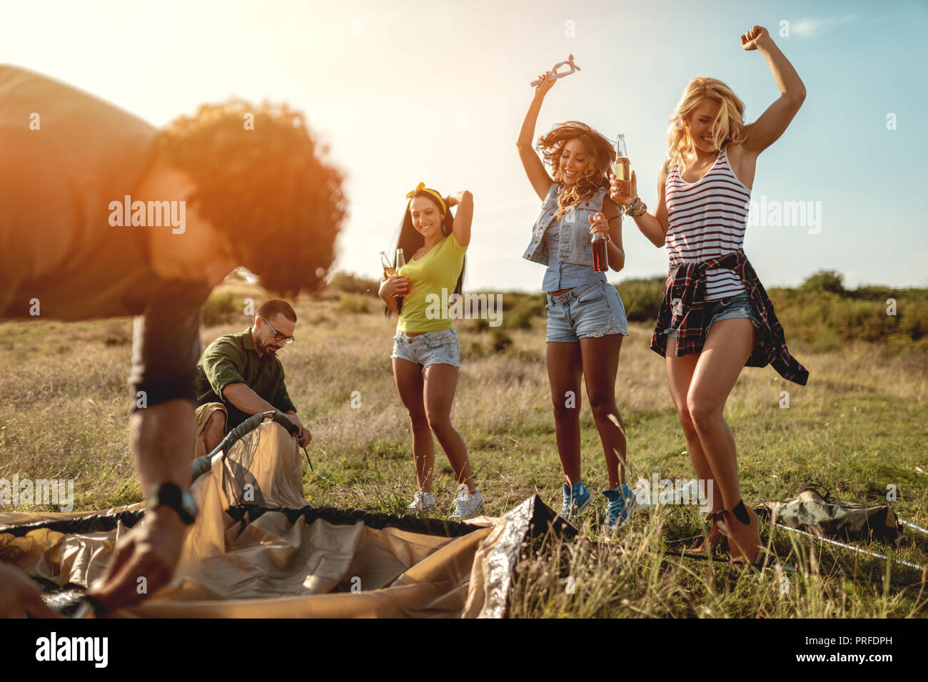 The young happy friends are preparing for camping. They're installing a tent on a suitable place in a meadow and their girlfriends are offering the be Stock Photo