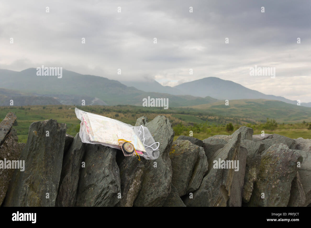 Ordnance Survey map in a plastic case with a compass on a drystone wall in the Lake District, overlooking a cloudy and misty Broughton Moor in Furness. Stock Photo