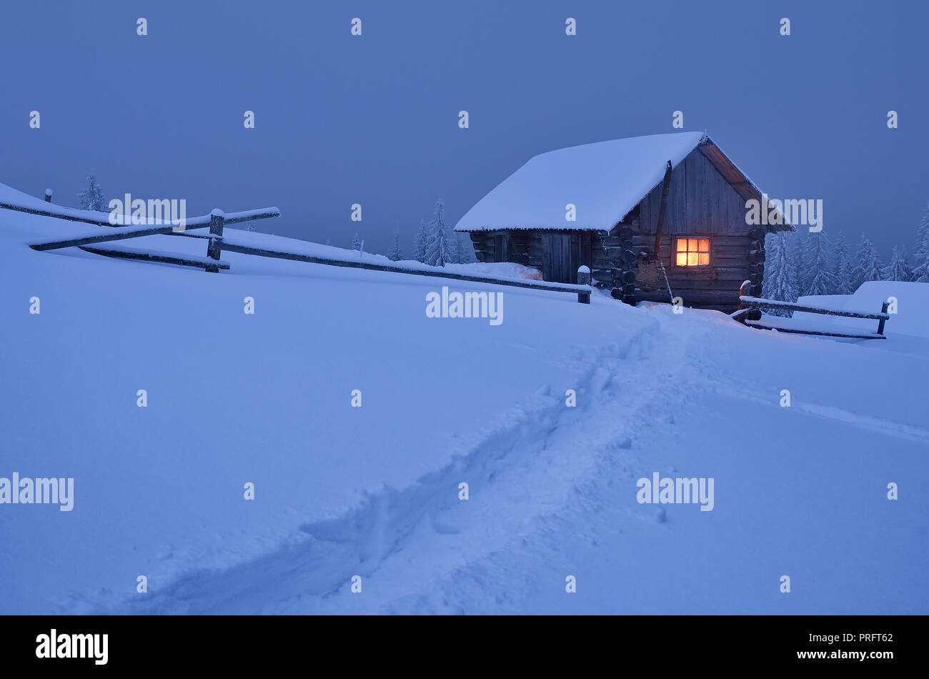 Winter landscape at night. Wooden house with a light in the window. The trail in the snow. Christmas view Stock Photo