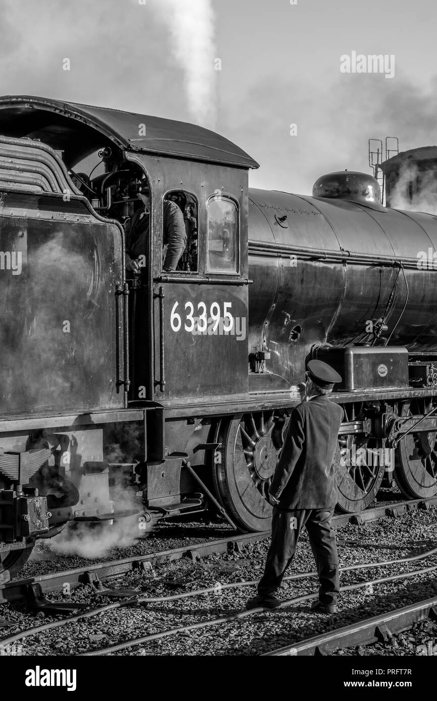 Black & white close up of vintage UK steam locomotive on track at heritage railway station. Train guard speaks to steam train crew in cab. Stock Photo