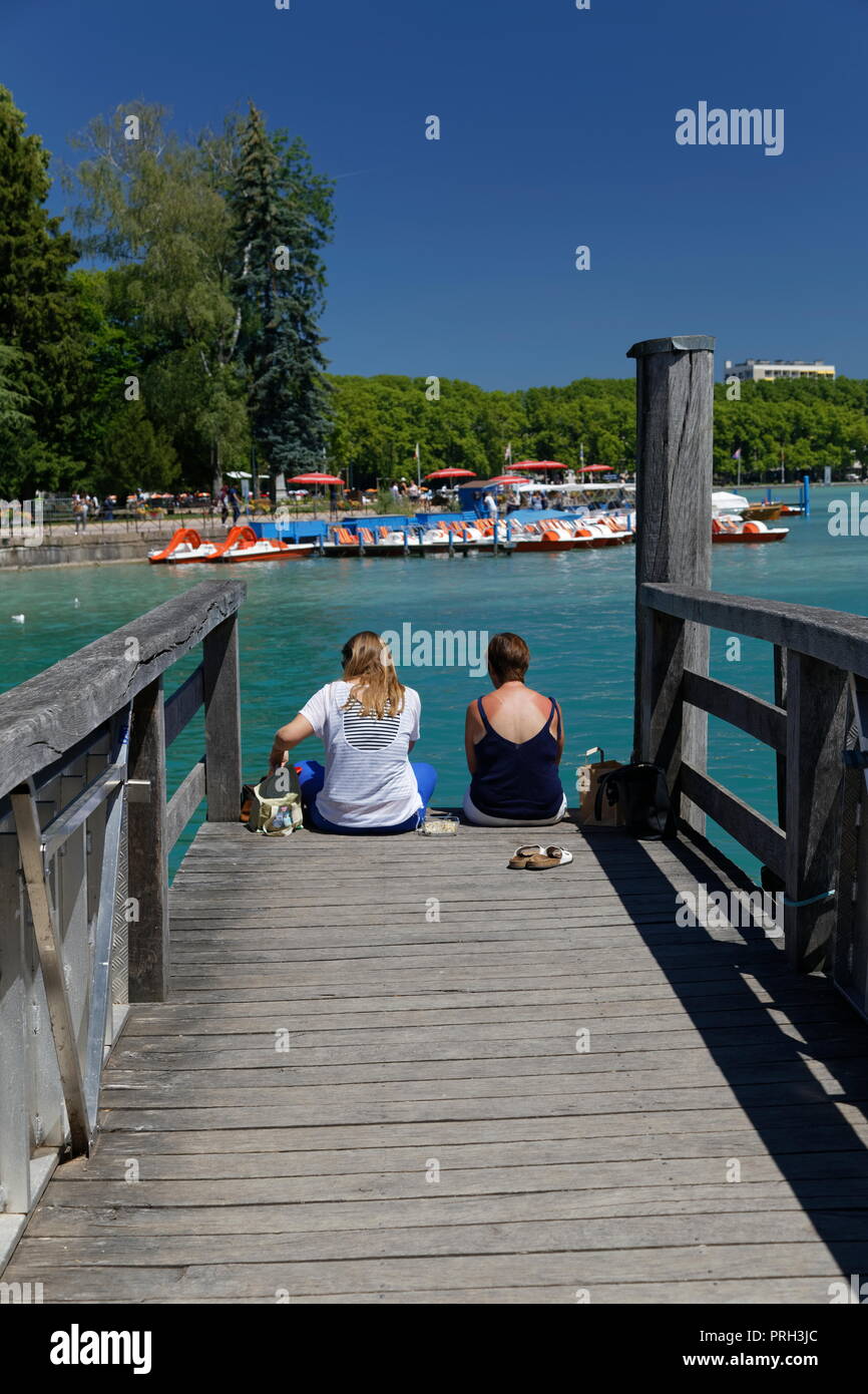 Two females sitting on the end of a wooden jetty extending out to the crystal clear waters of Lake Annecy France Stock Photo
