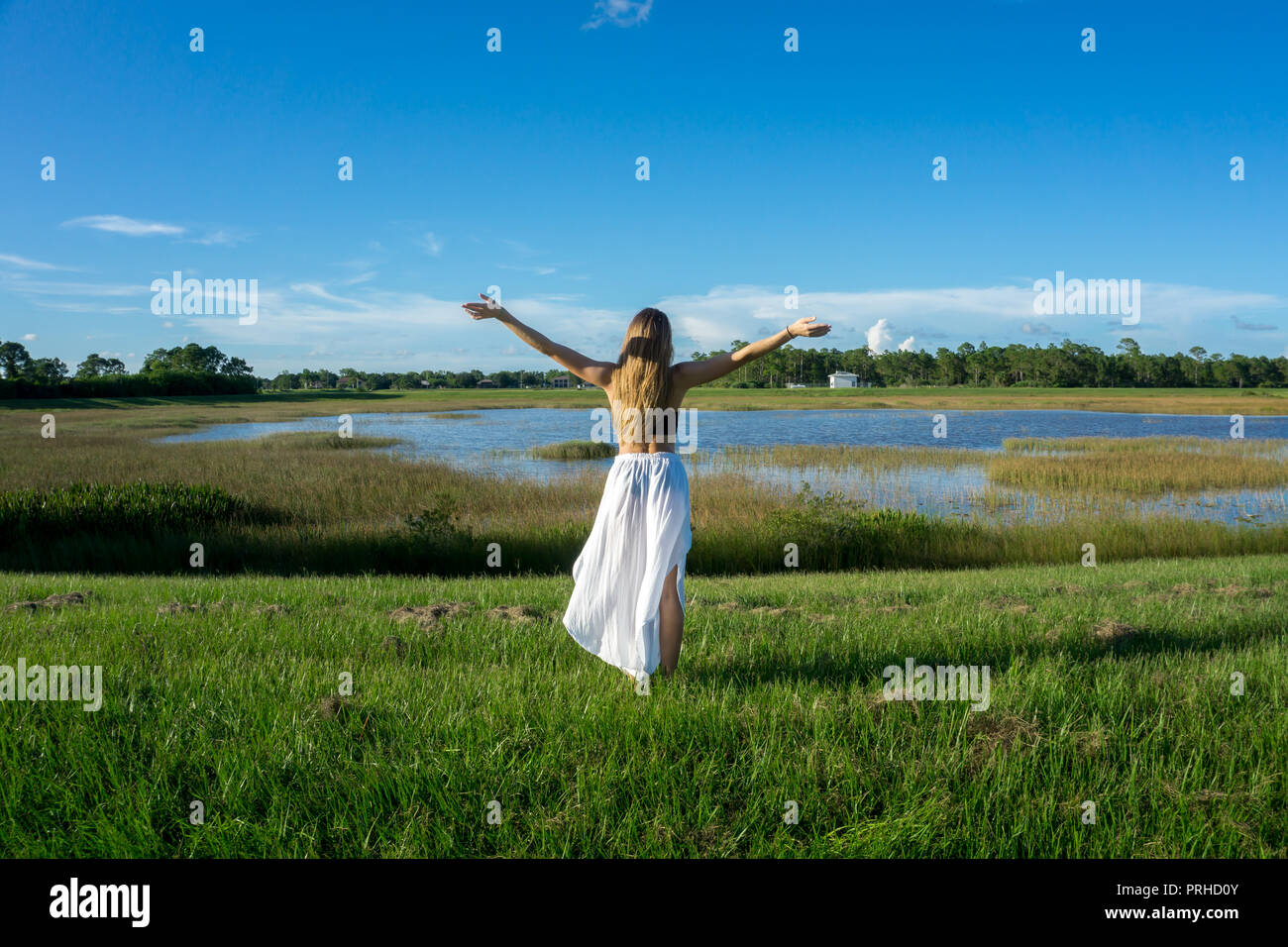 Blonde young woman standing backwards in a beautiful field landscape outdoors with raise hands arms to the sky Stock Photo