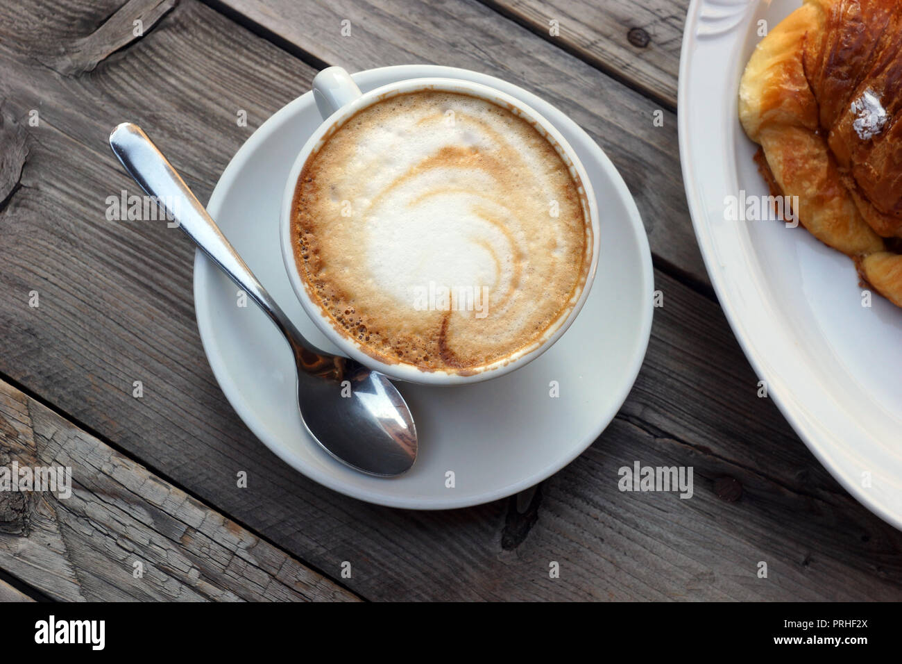 Wonderful traditional Italian breakfast. Delicious hot Italian cappuccino stands in a cup with a saucer on a wooden table. Сroissant on a plate. Stock Photo