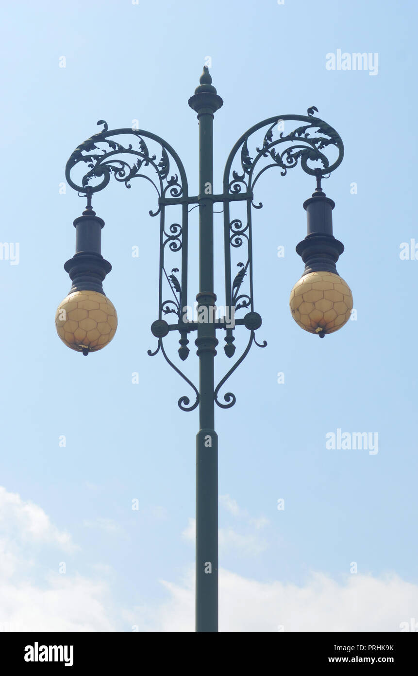 A wrought-iron lamp post with leaf details against a blue sky. The lamps are yellow with a pentagon pattern. Stock Photo