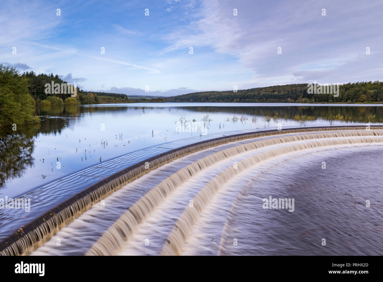 Water flowing over spillway from calm scenic tree-lined lake, under deep blue sky - Fewston Reservoir, Washburn Valley, North Yorkshire, England, UK. Stock Photo