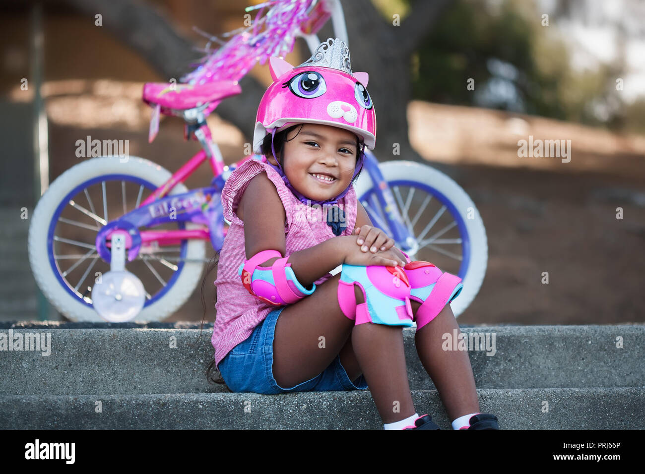 Pretty young girl with bicycle safety gear smiling and sitting in front of her new bicycle with training wheels in an outdoor park Stock Photo